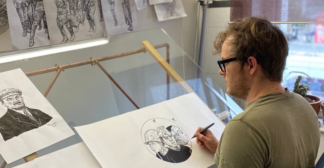 Peter Mammes drawing at a glass-topped studio desk surrounded by figurative studies.